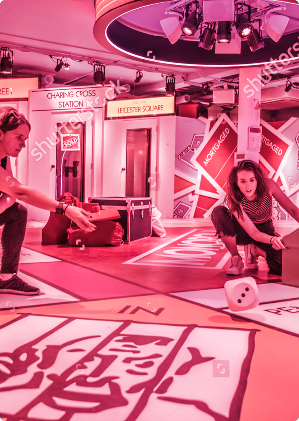 People playing a giant Monopoly board game indoors with oversized game pieces and dice.