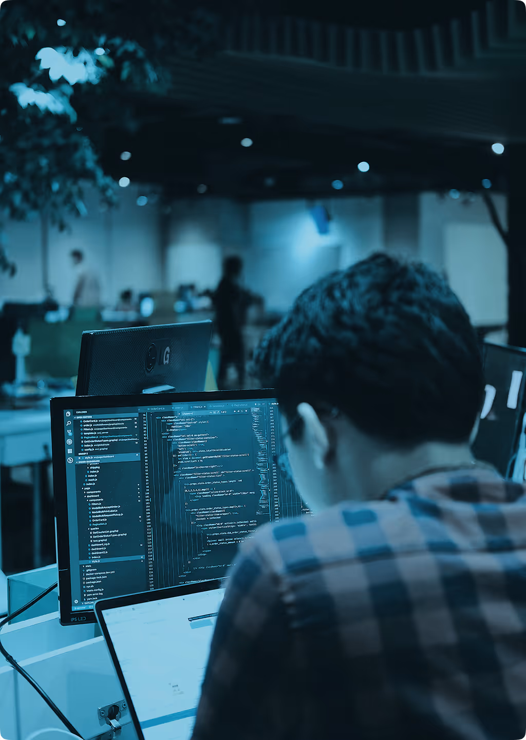 Man in a red and blue checkered shirt coding on a computer with dual monitors in an office.
