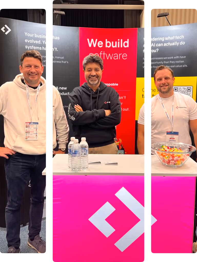Three men smiling at a software booth with a pink logo and a red sign saying 'We build software'.
