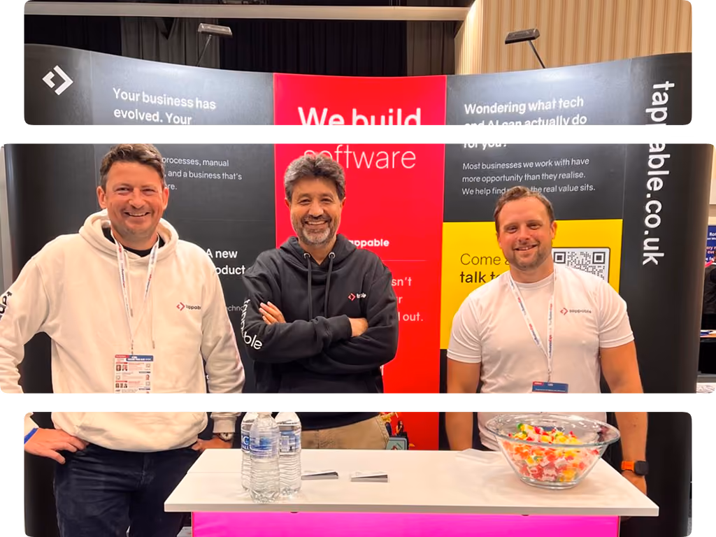 Three men smiling at a software booth with a pink logo and a red sign saying 'We build software'.