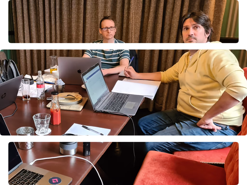 Two men sitting at a table with laptops, notebooks, and drinks, engaged in work or discussion.