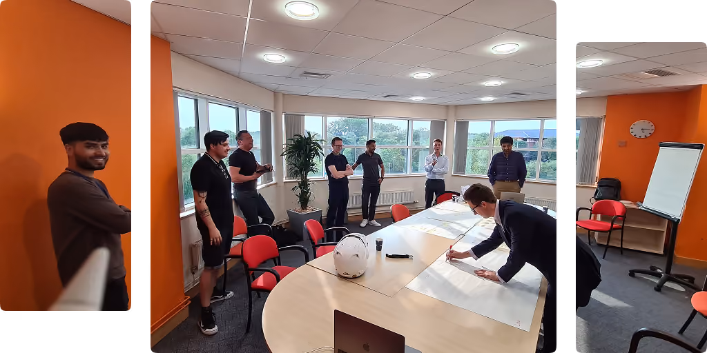 Eight men in a meeting room, one writing on large sheets on the table while others watch.