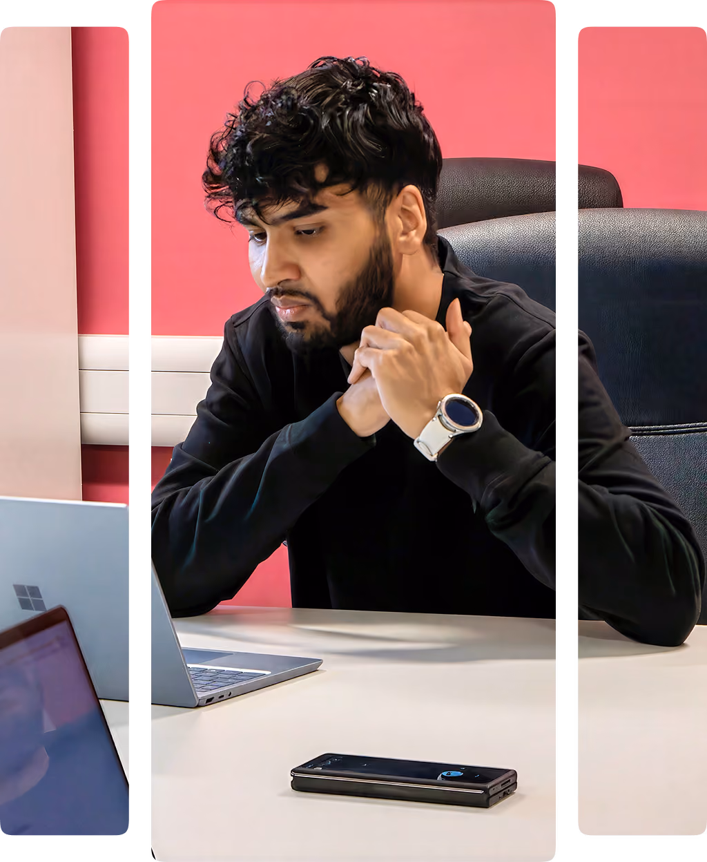 Man with curly hair and beard sitting at a desk, looking at a laptop, with smartphone on table.