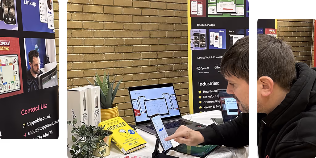Man using a smartphone on stand at a booth with laptops, books, and promotional banners.