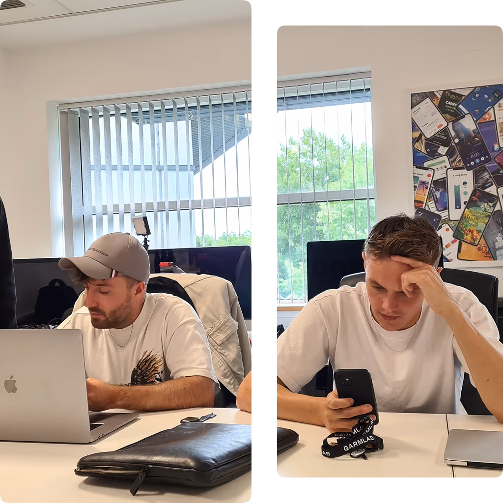 Two men at a table in an office, one working on a laptop, the other looking at a phone.