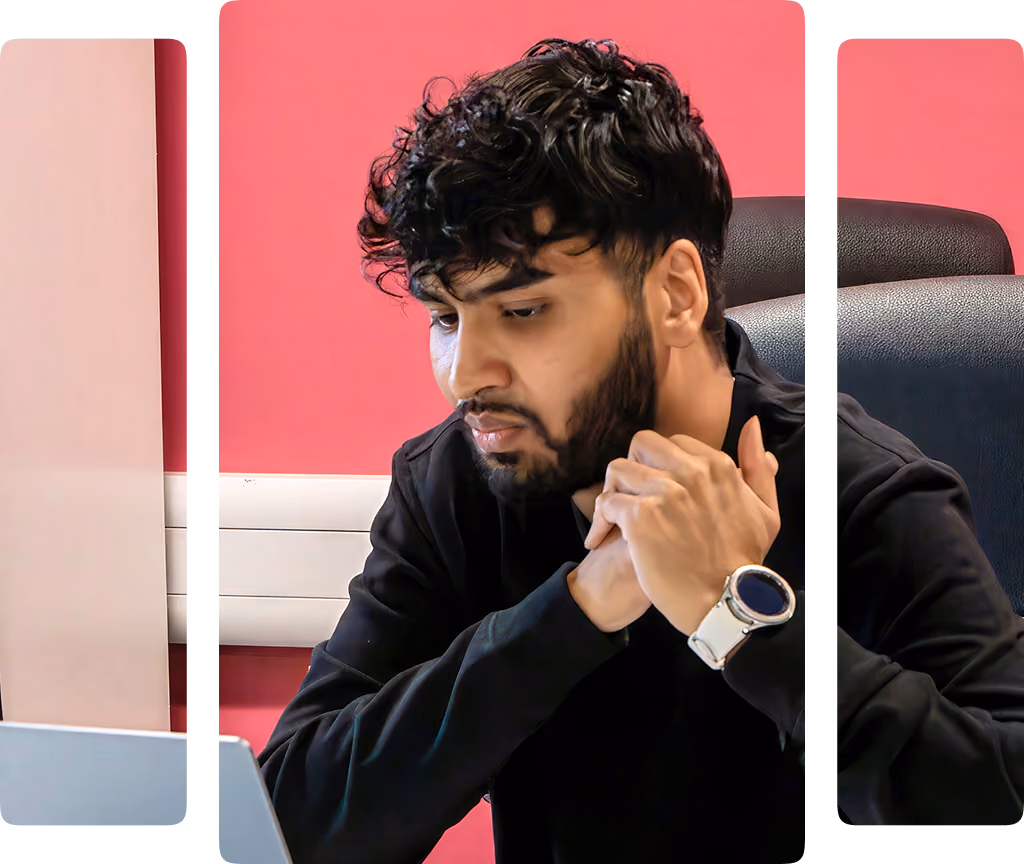 Man with curly hair and beard sitting at a desk, looking at a laptop, with smartphone on table.