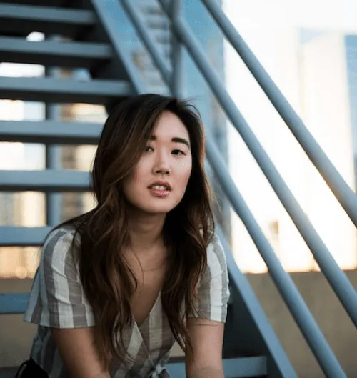 Person with long hair sitting on outdoor stairs with a cityscape in the background.