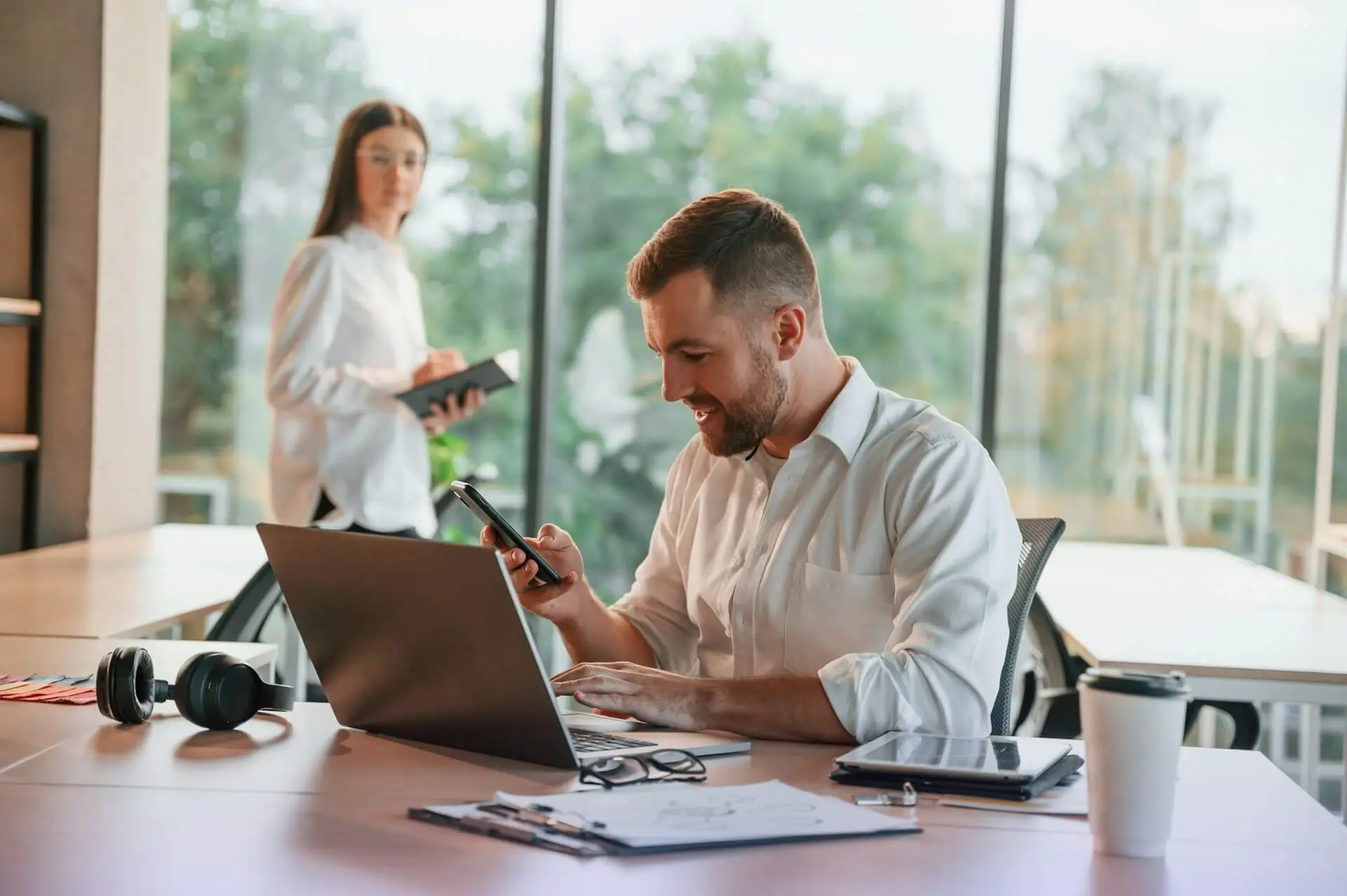 Man using smartphone and laptop at a desk in a modern office setting