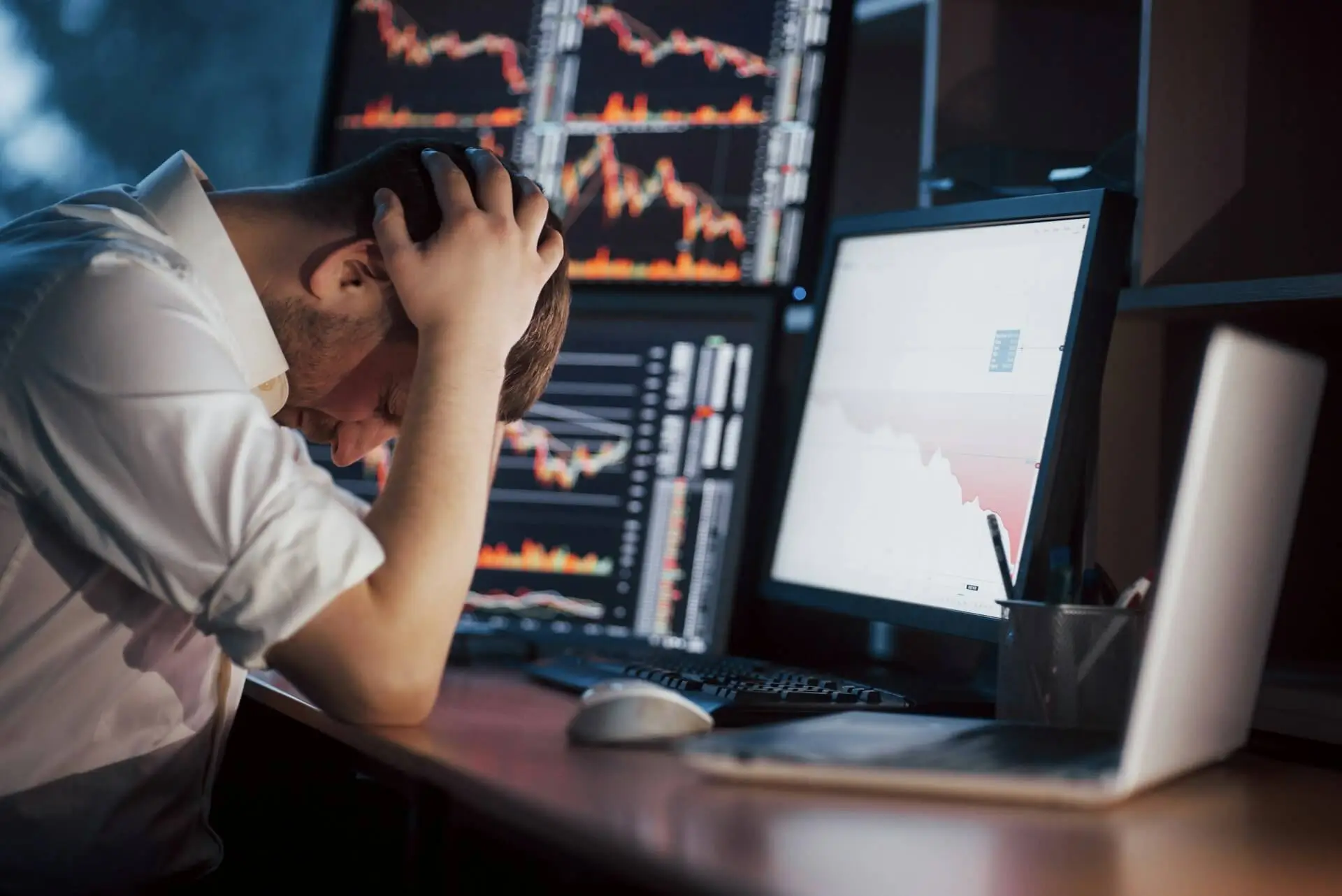 Man sitting at a desk holding his head in frustration while looking at multiple computer screens showing falling graphs and data loss