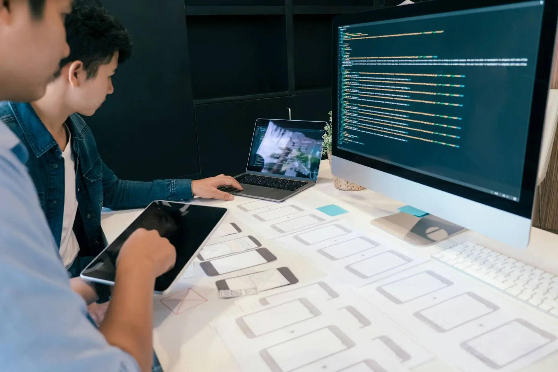 Two professionals reviewing mobile app wireframes while coding on a desktop and laptop