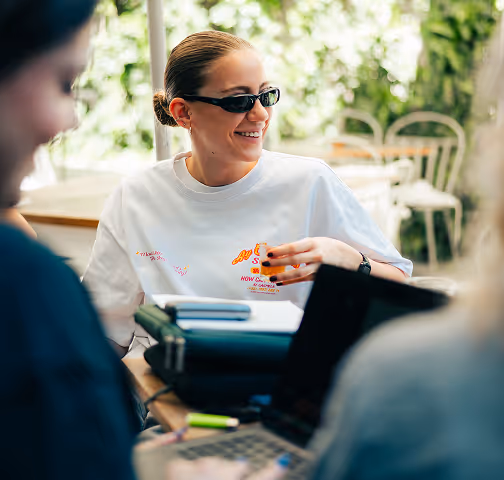 Young woman in sunglasses smiling and sitting at a table with laptops and notebooks outdoors.