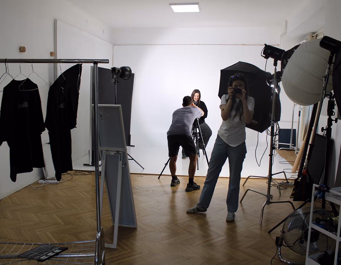 Photography studio scene with two people taking photos of a seated woman against a white backdrop.