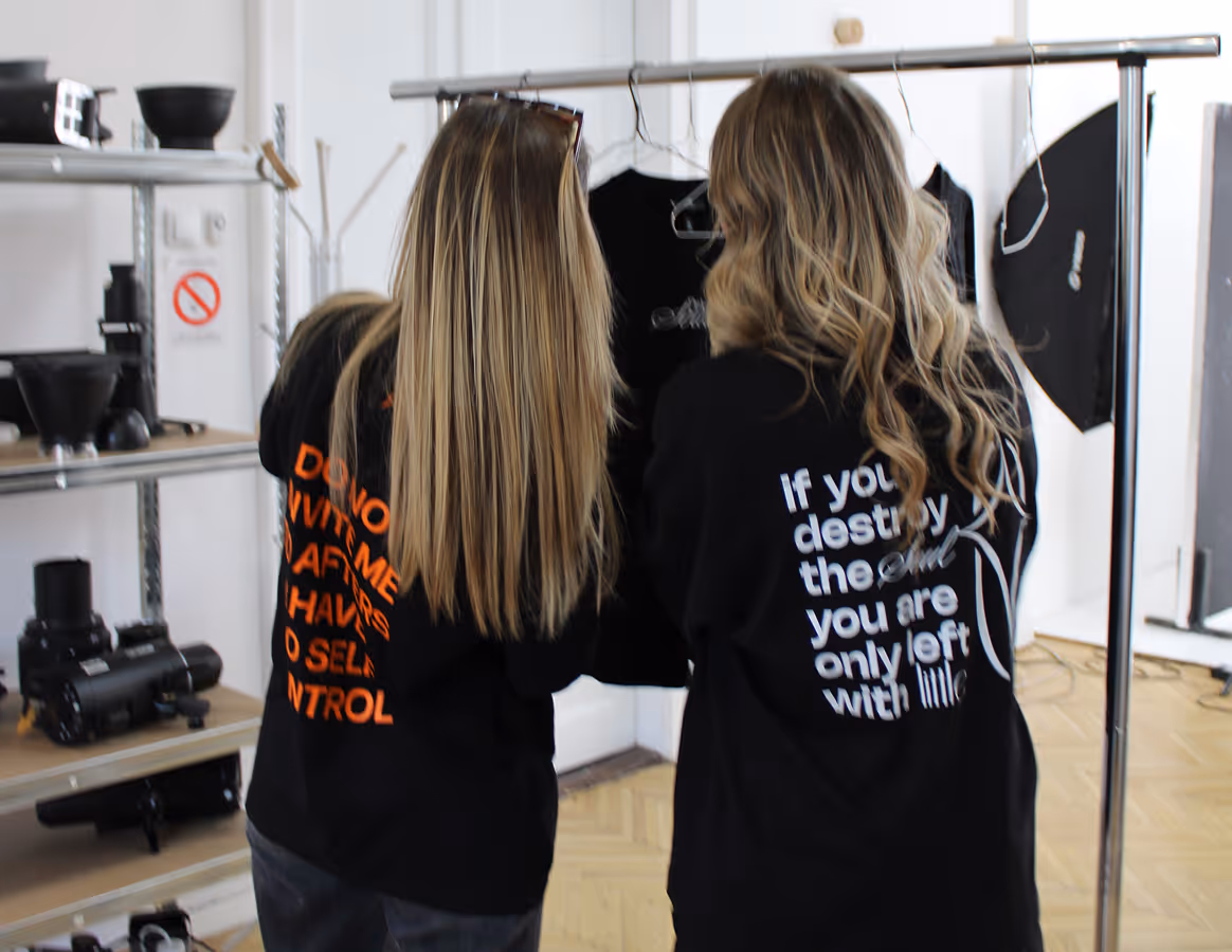 Two women seen from behind wear black shirts with text, standing near a clothing rack in a studio.