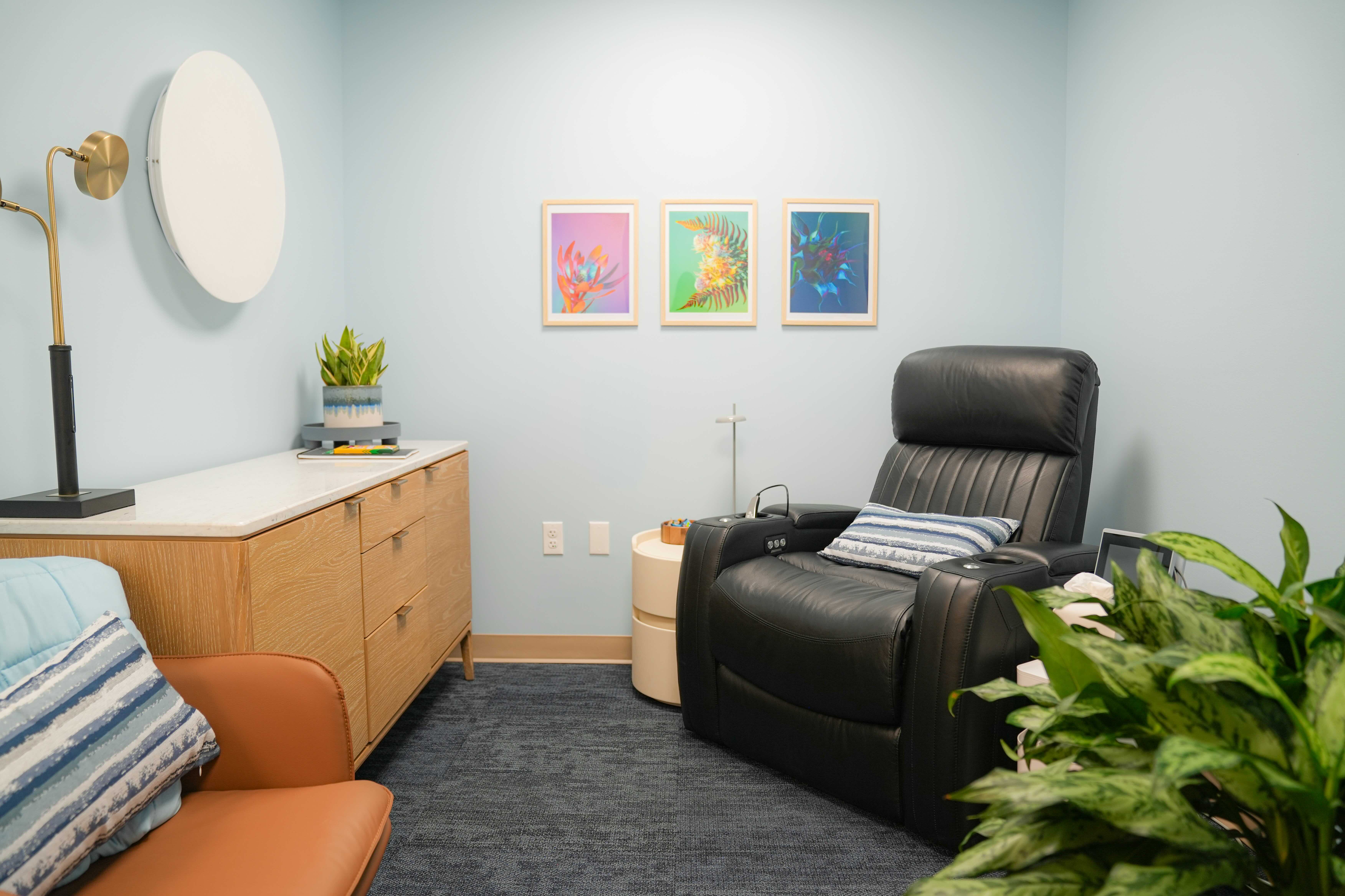 A serene treatment room with a black recliner, warm chair, plant, and colorful framed botanical art