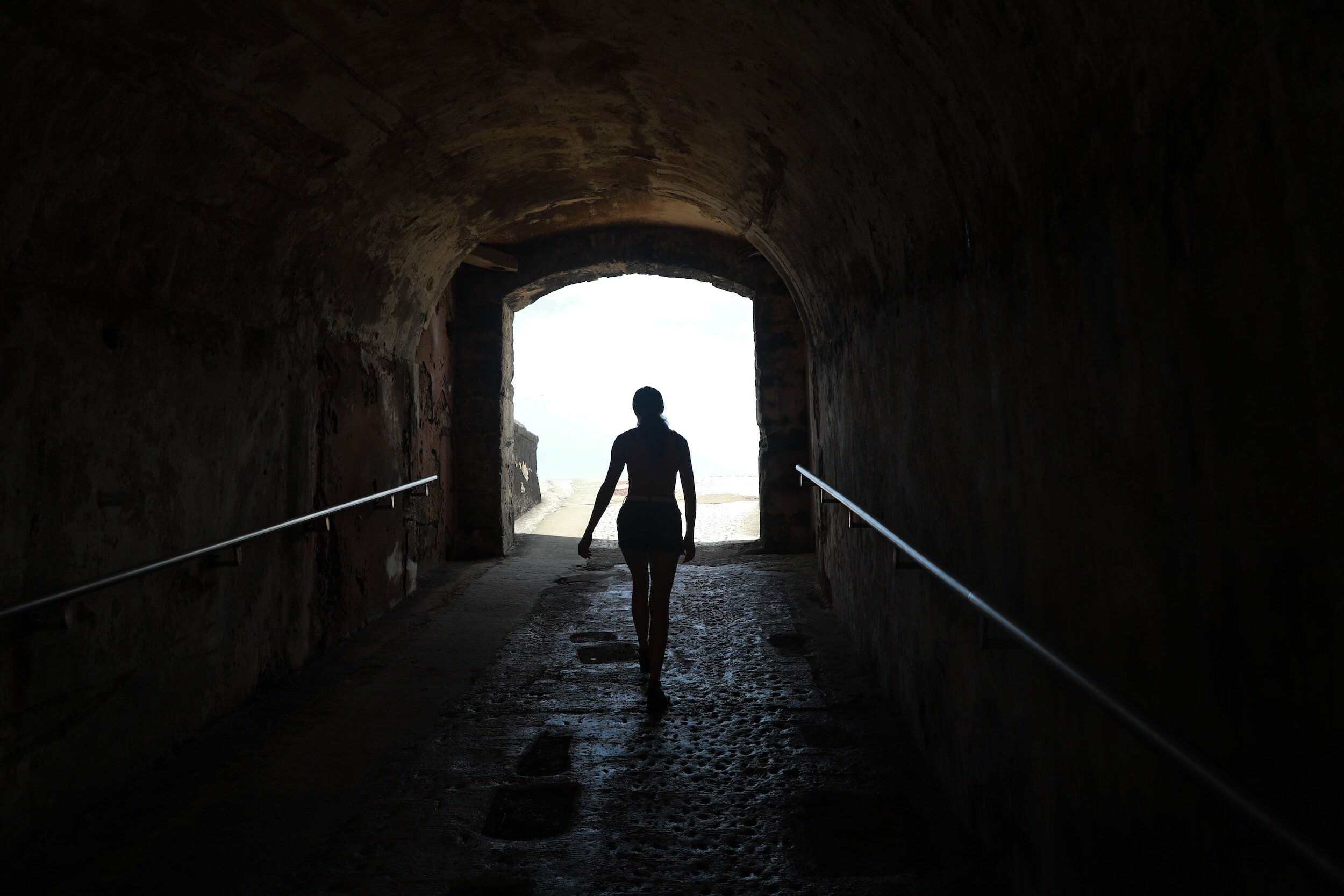 Silhouette of a person walking through a dark stone tunnel toward bright daylight