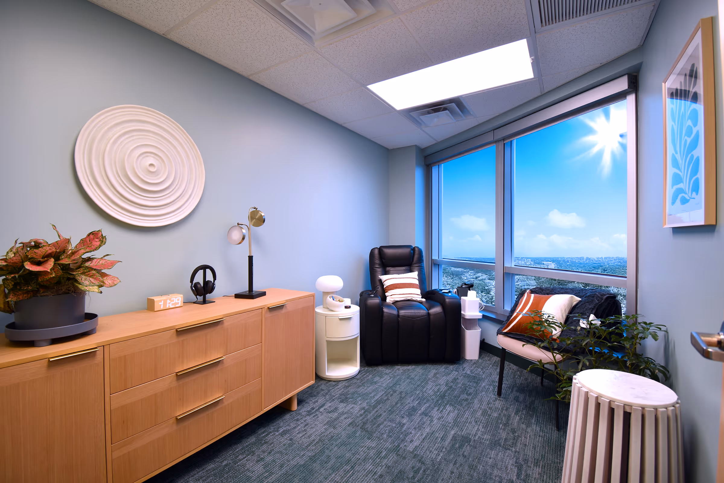 A calm Lumin Health treatment room with a recliner, wood console, spiral wall art, and city view