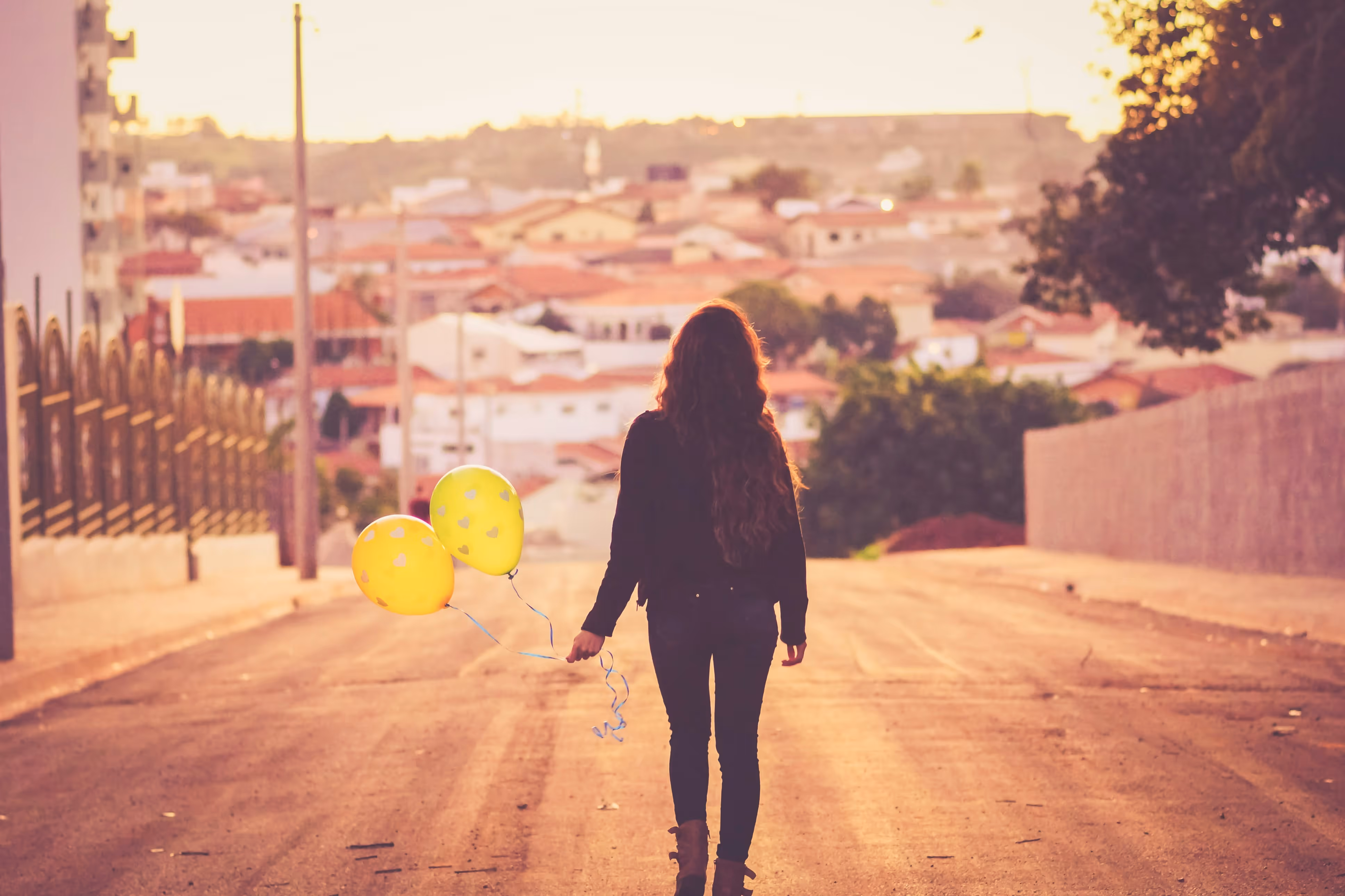 A woman with long hair walks down a sunlit street holding two yellow balloons, town rooftops ahead