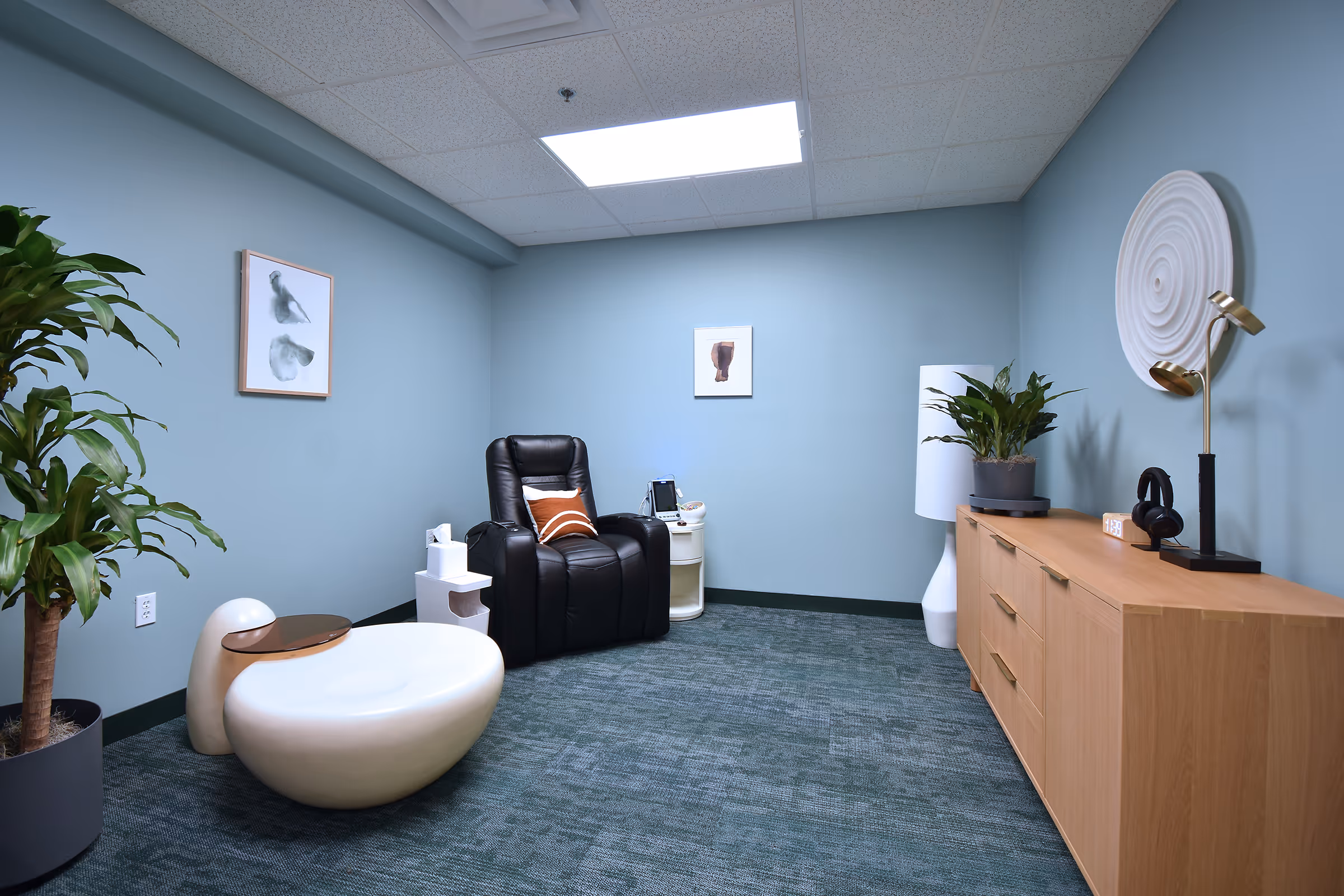 A calm treatment room with a black recliner, soft lighting, plants, and a spiral wall sculpture