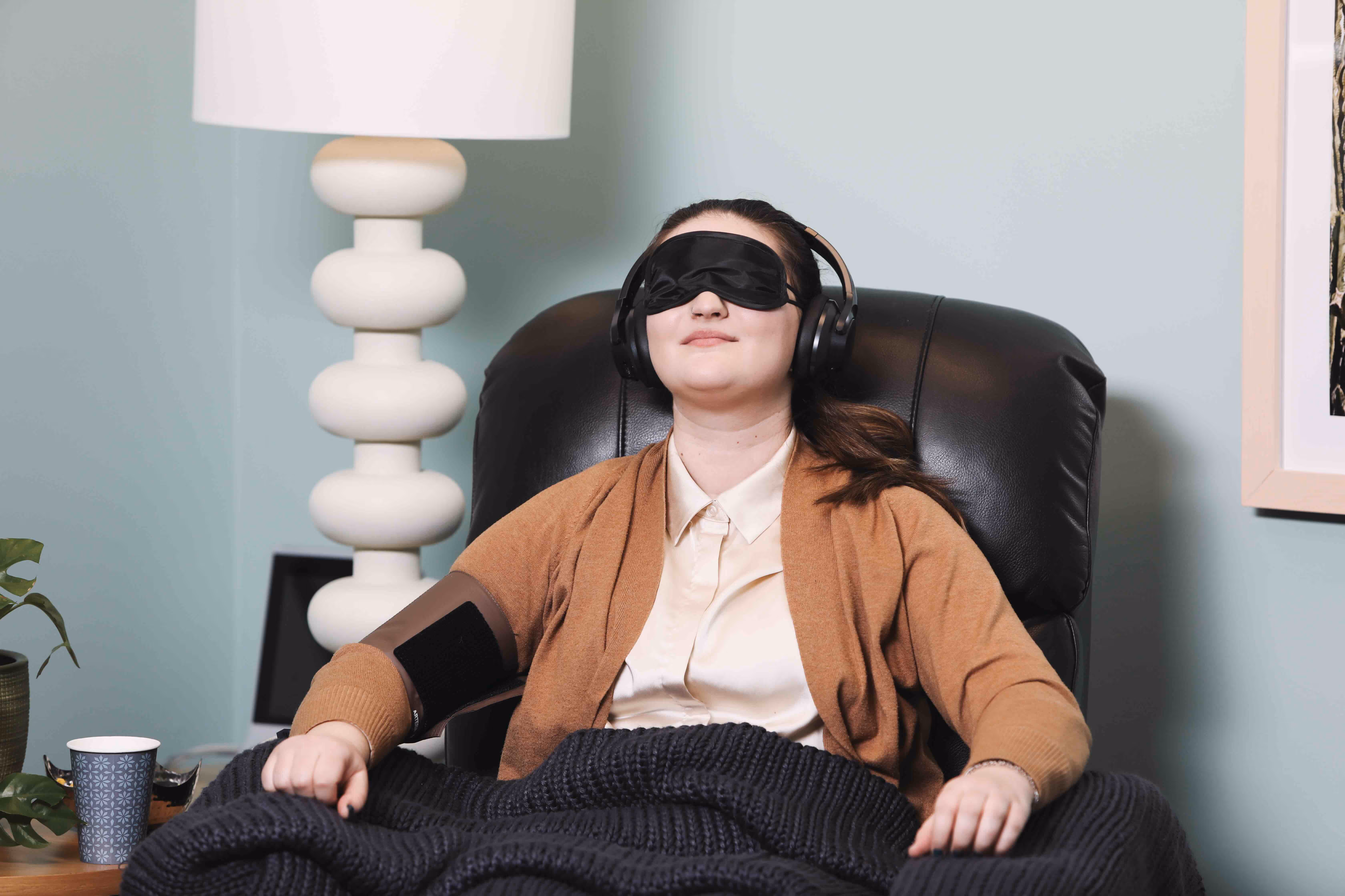 A woman relaxing in a leather recliner with an eye mask, headphones, and a blanket in a calm, warmly lit room