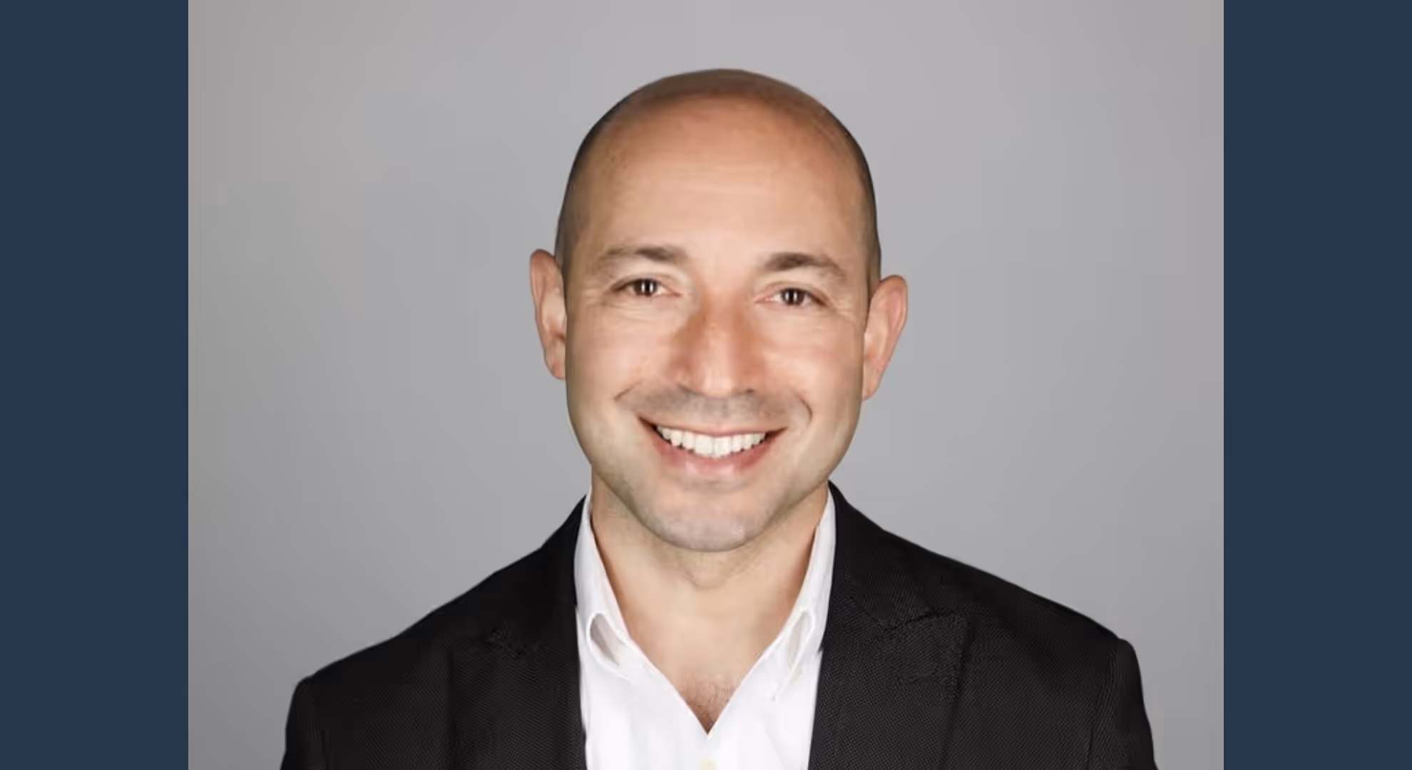 Smiling man in a black blazer and white shirt, professional headshot on gray backdrop