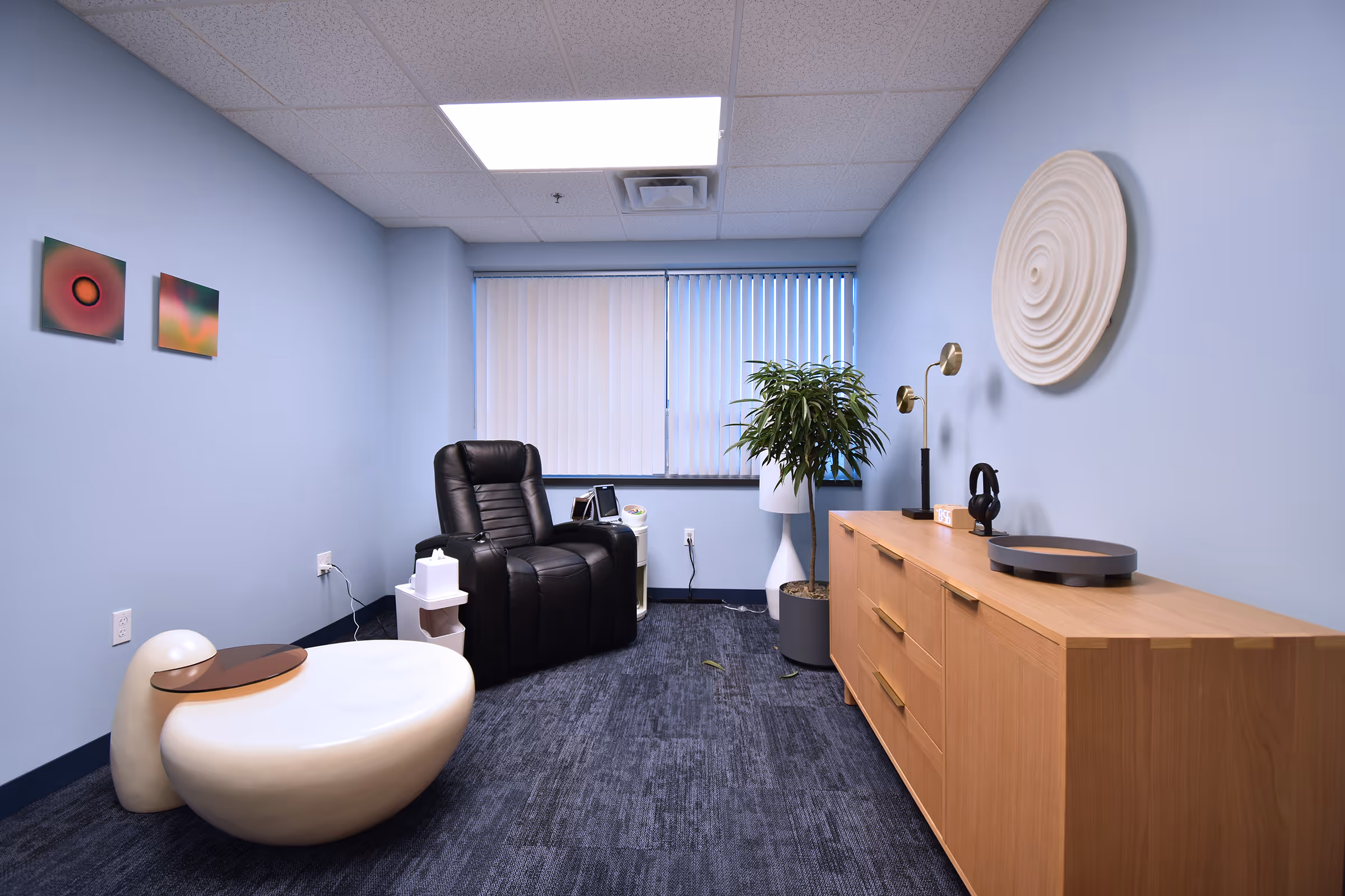 A calm treatment room with a black recliner, soft blue walls, a potted plant, and a spiral wall sculpture