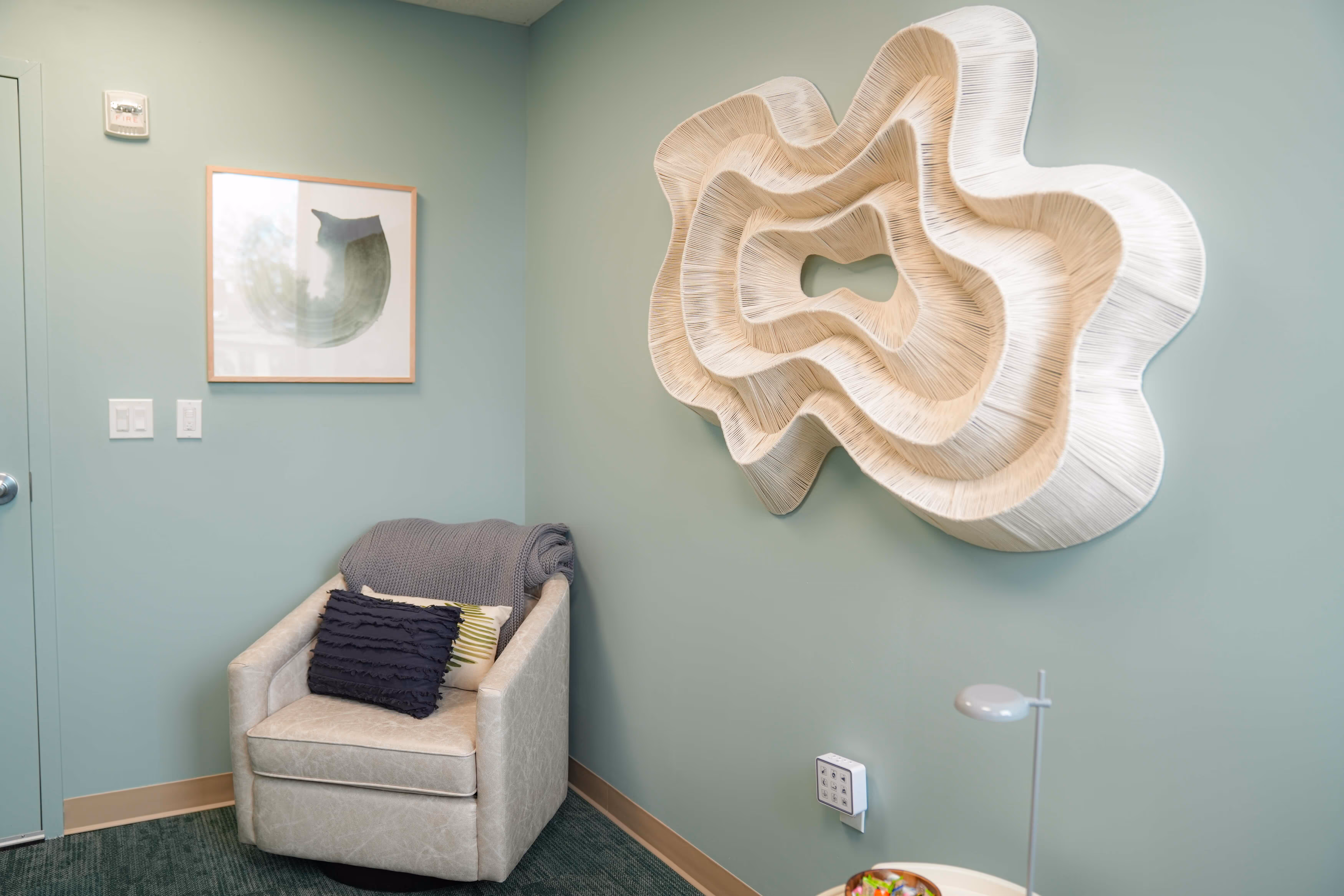 A serene treatment room with a soft armchair, folded blanket, and a sculptural wood wall piece on green walls