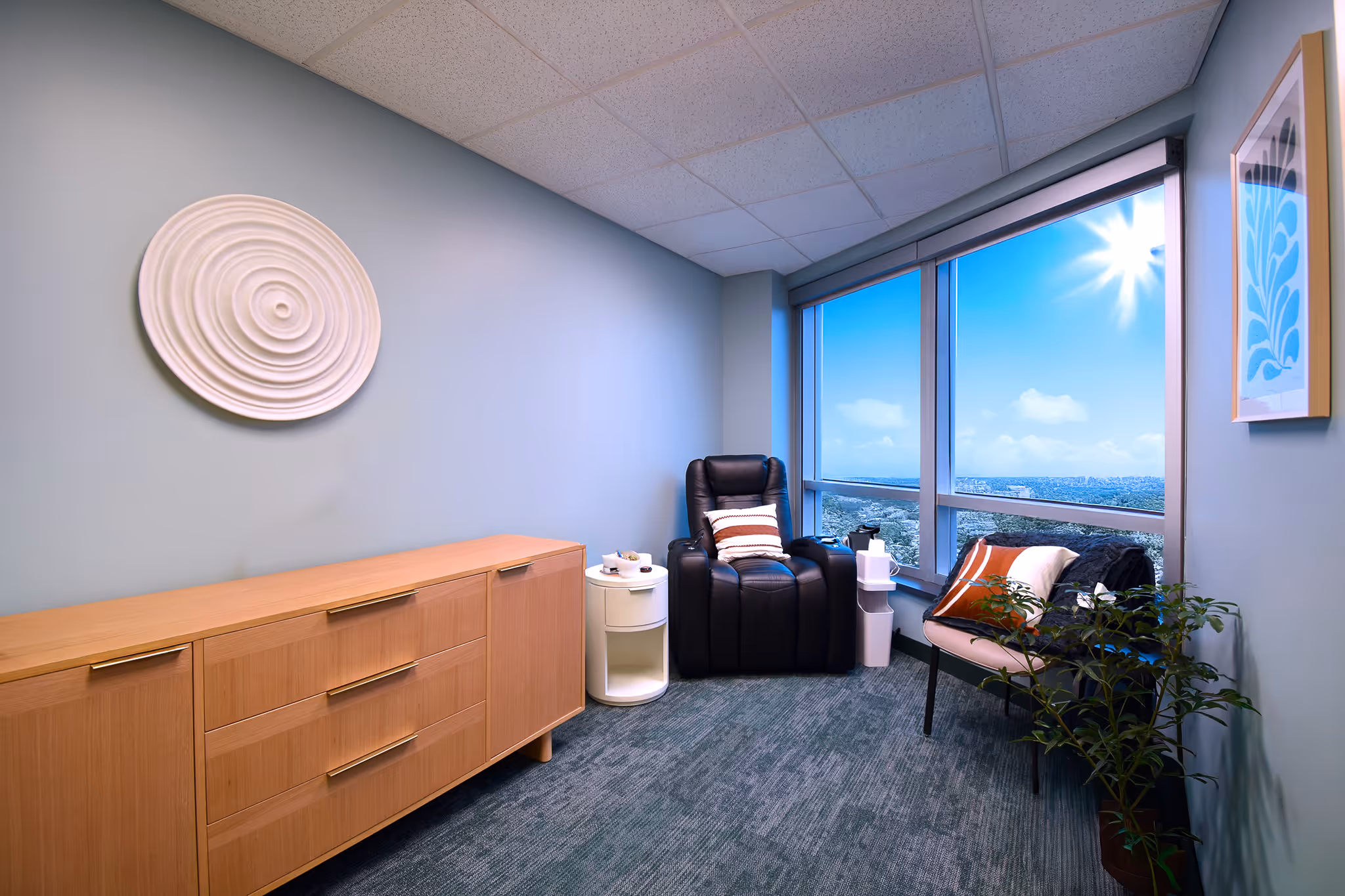 A bright treatment room with a recliner and bench beside a sunlit window overlooking a city skyline