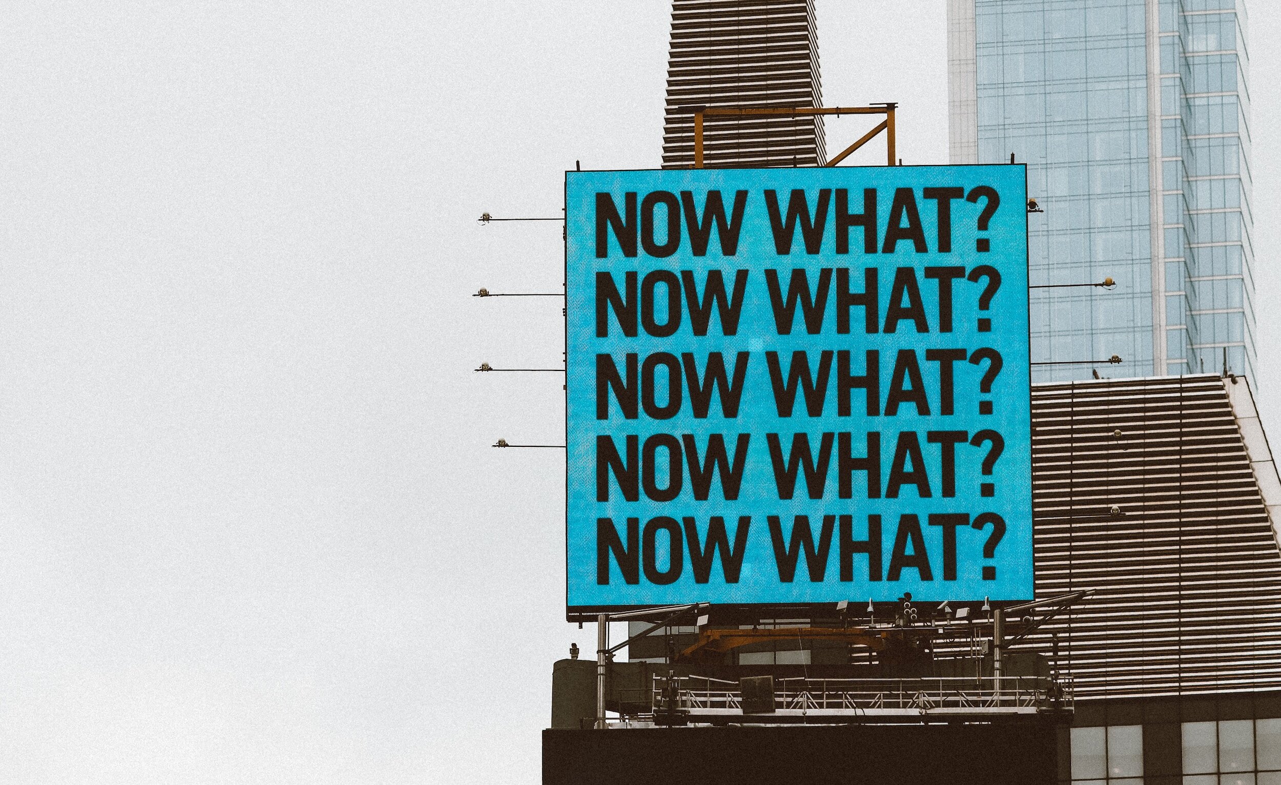 A blue billboard repeating the question 'Now what?' against a cloudy city skyline
