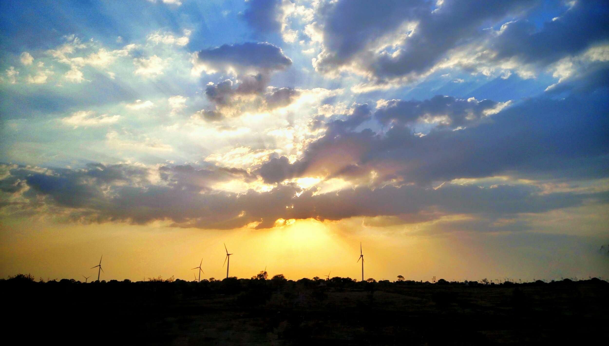 Sun breaking through dramatic clouds over a plain dotted with distant wind turbines at dusk