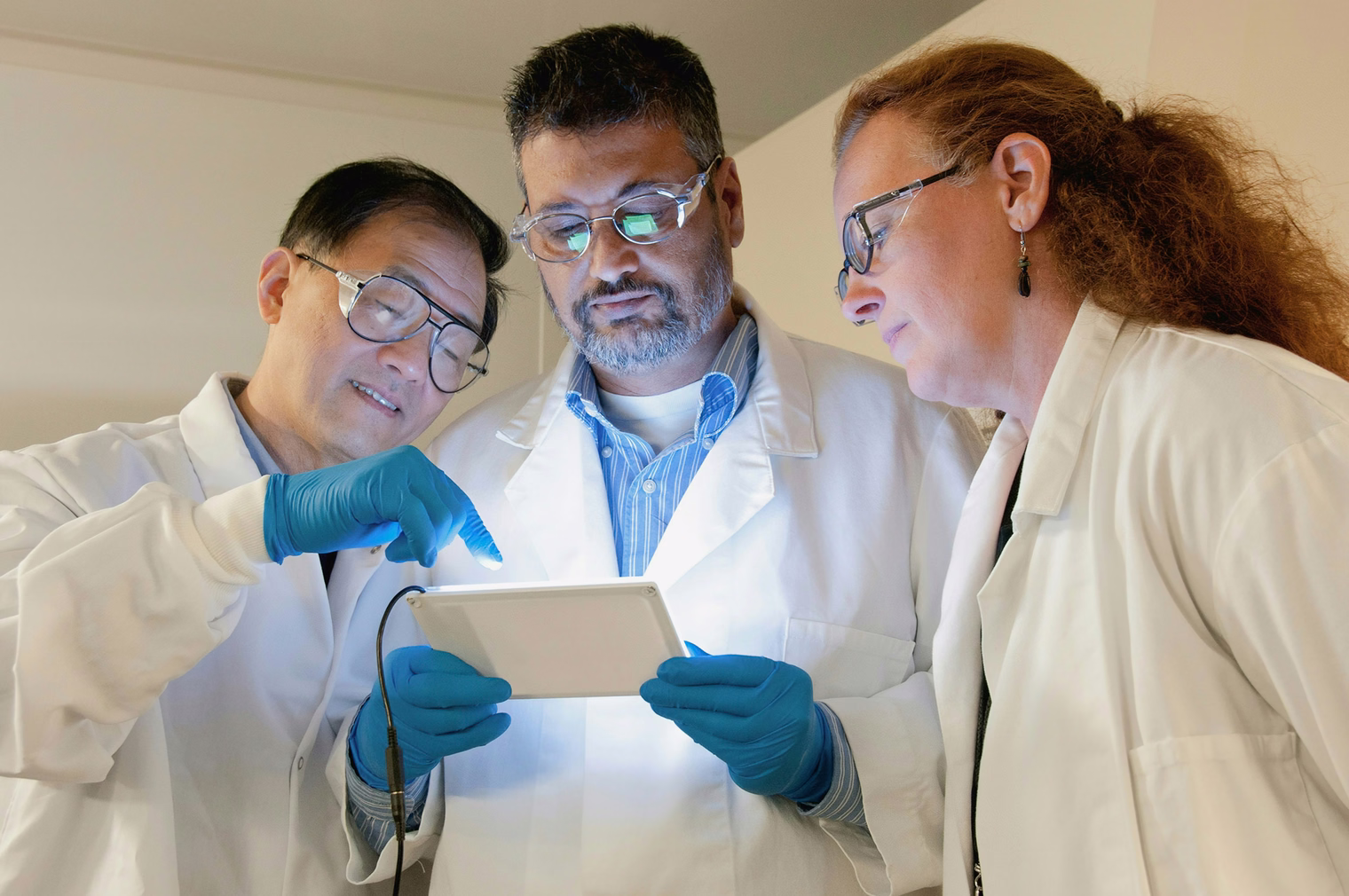 Three clinicians in white coats and gloves studying a tablet together