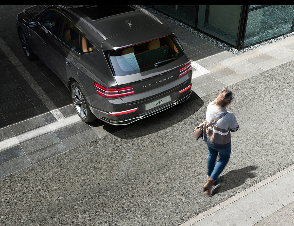 Storr Green GV80 backing out of a parking lot, and a woman is passing behind the vehicle.