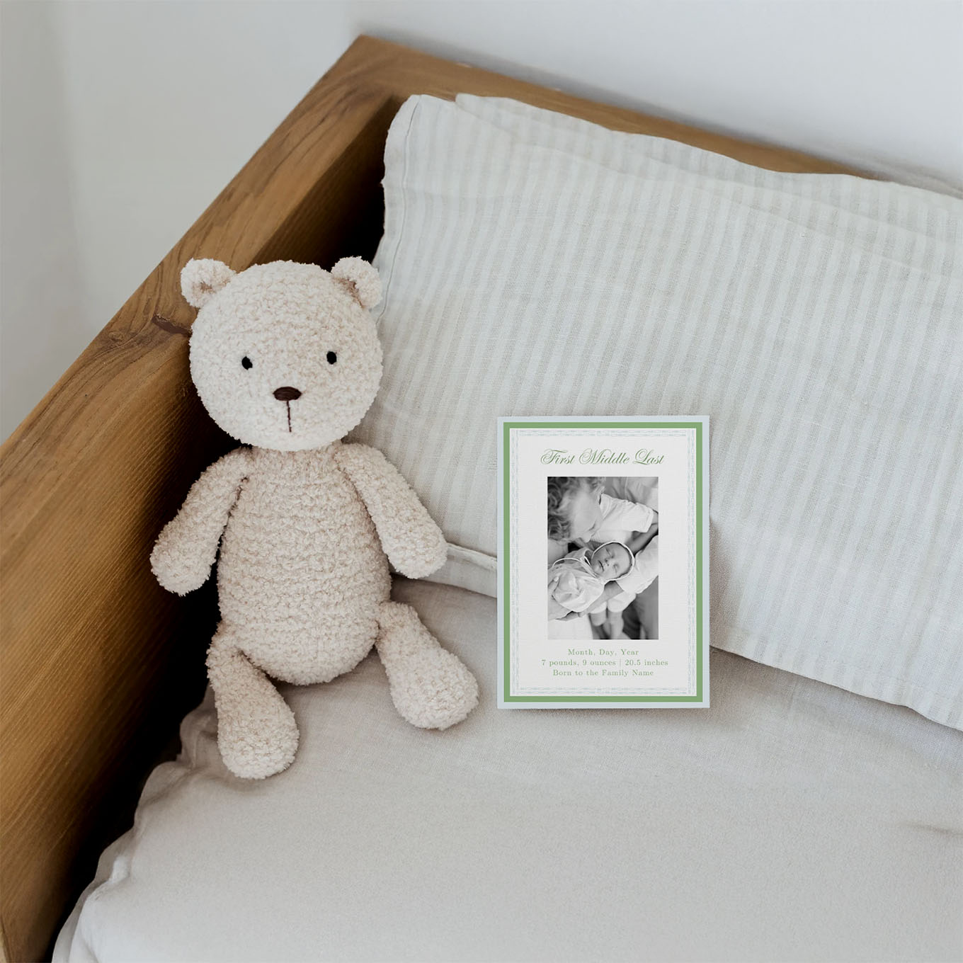 Soft beige teddy bear sitting on a bed next to a baby announcement card featuring a photo of a newborn with an older child.