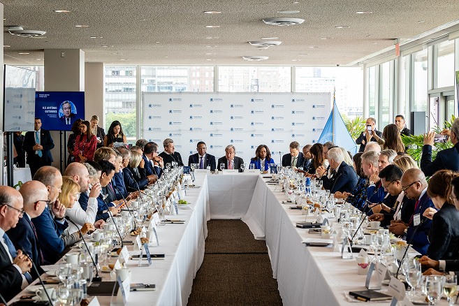 Large group of formally dressed people seated around a U-shaped conference table in a bright room, engaged in a meeting with a backdrop and flags behind the head table.