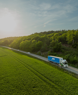White truck with a blue trailer driving on a road beside green fields and forest under a cloudy sky.