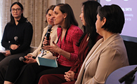 Five women seated in a panel discussion, one woman in red speaking into a microphone.