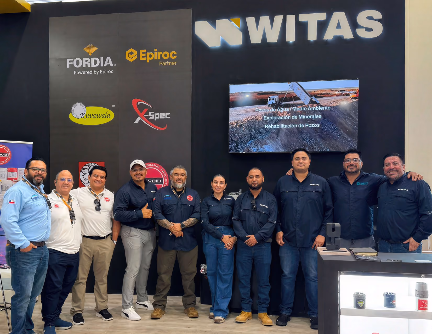 Tres personas posan detrás de un mostrador con piezas industriales expuestas y un letrero que dice FORDIA Powered by Epiroc.