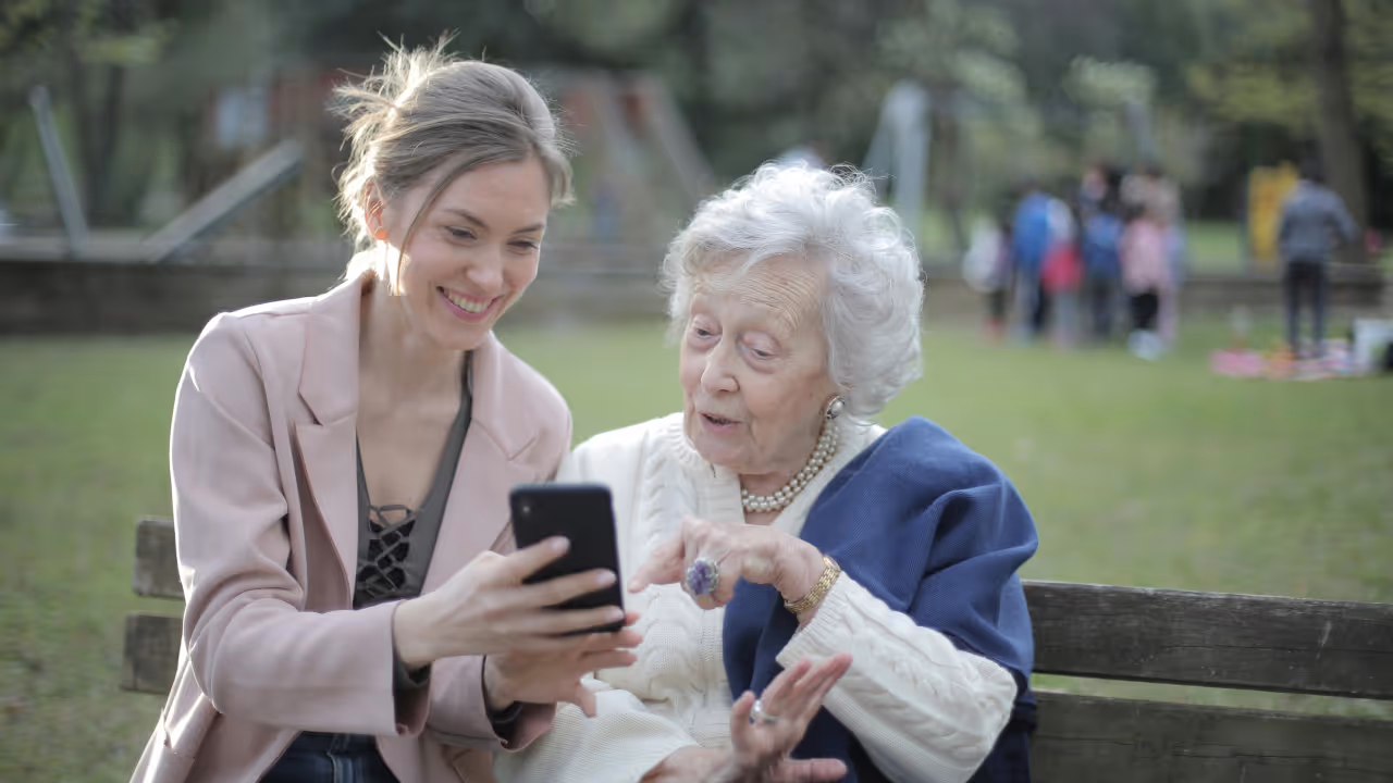 A young woman in a pink blazer sits on a park bench with an elderly woman wearing a white sweater and navy cardigan. They are both smiling while looking at and interacting with a smartphone together. In the background, there is a grassy area with other people visible.