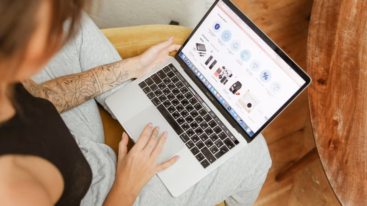 An overhead view of someone using a MacBook laptop while sitting on what appears to be a couch or chair with gray cushioning. The person has a visible floral tattoo on their arm and is wearing a dark colored top. On the laptop screen, an e-commerce or shopping website is displayed showing various product icons and images.