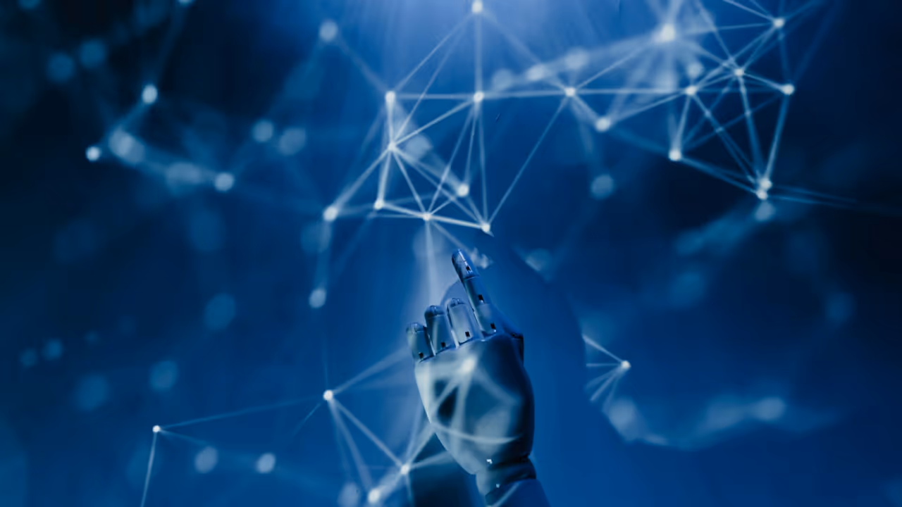 A robotic hand with metallic fingers reaching out toward a network of interconnected glowing nodes and lines, set against a dark blue background. The image conveys a sense of technology and artificial intelligence.