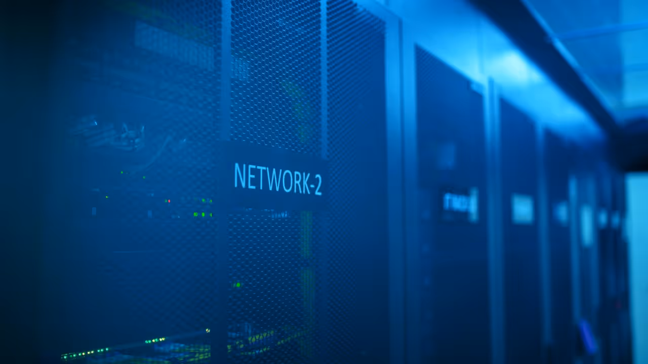 A row of server racks in a data centre, illuminated by blue lighting. The focus is on a rack labelled 'NETWORK-2', showing network equipment, cables, and indicator lights, representing a modern data storage and network management environment.