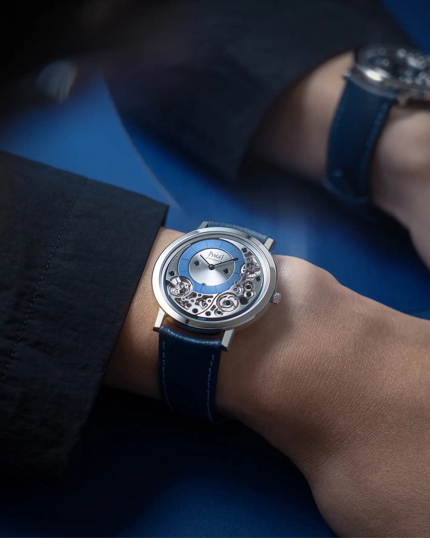 A Piaget Altiplano automatic watch with a blue dial and skeletonised movement worn on a wrist, photographed against a blue background.