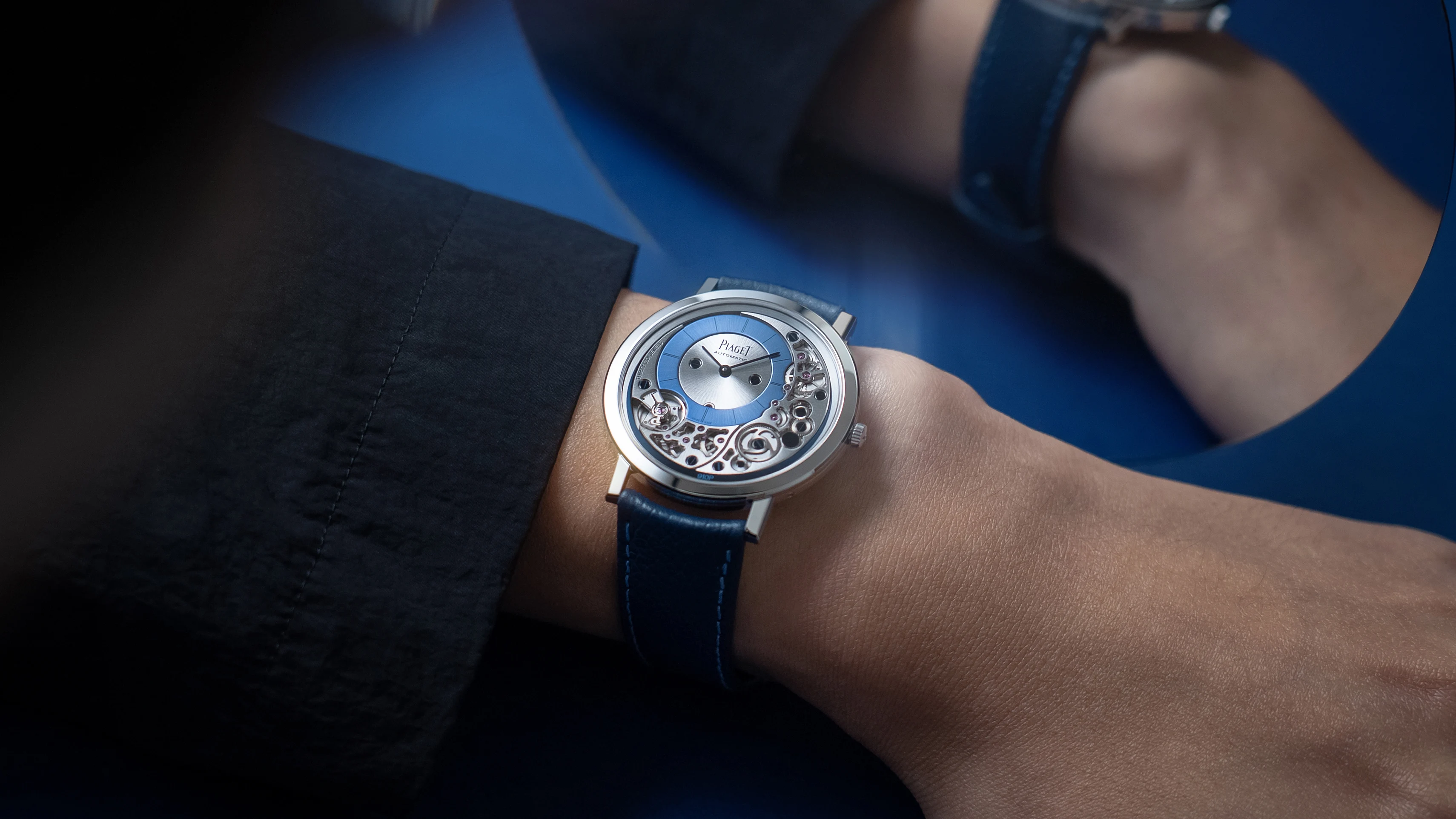 A Piaget Altiplano automatic watch with a blue dial and skeletonised movement worn on a wrist, photographed against a blue background.