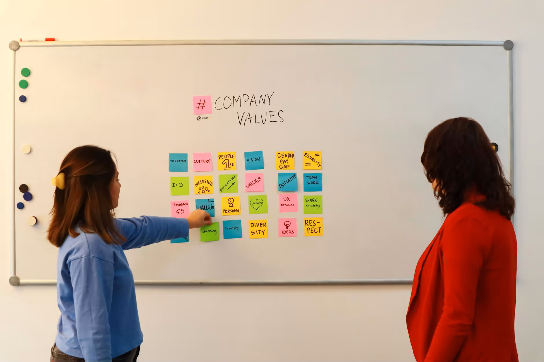 Two women in an office discussing company values on a whiteboard.