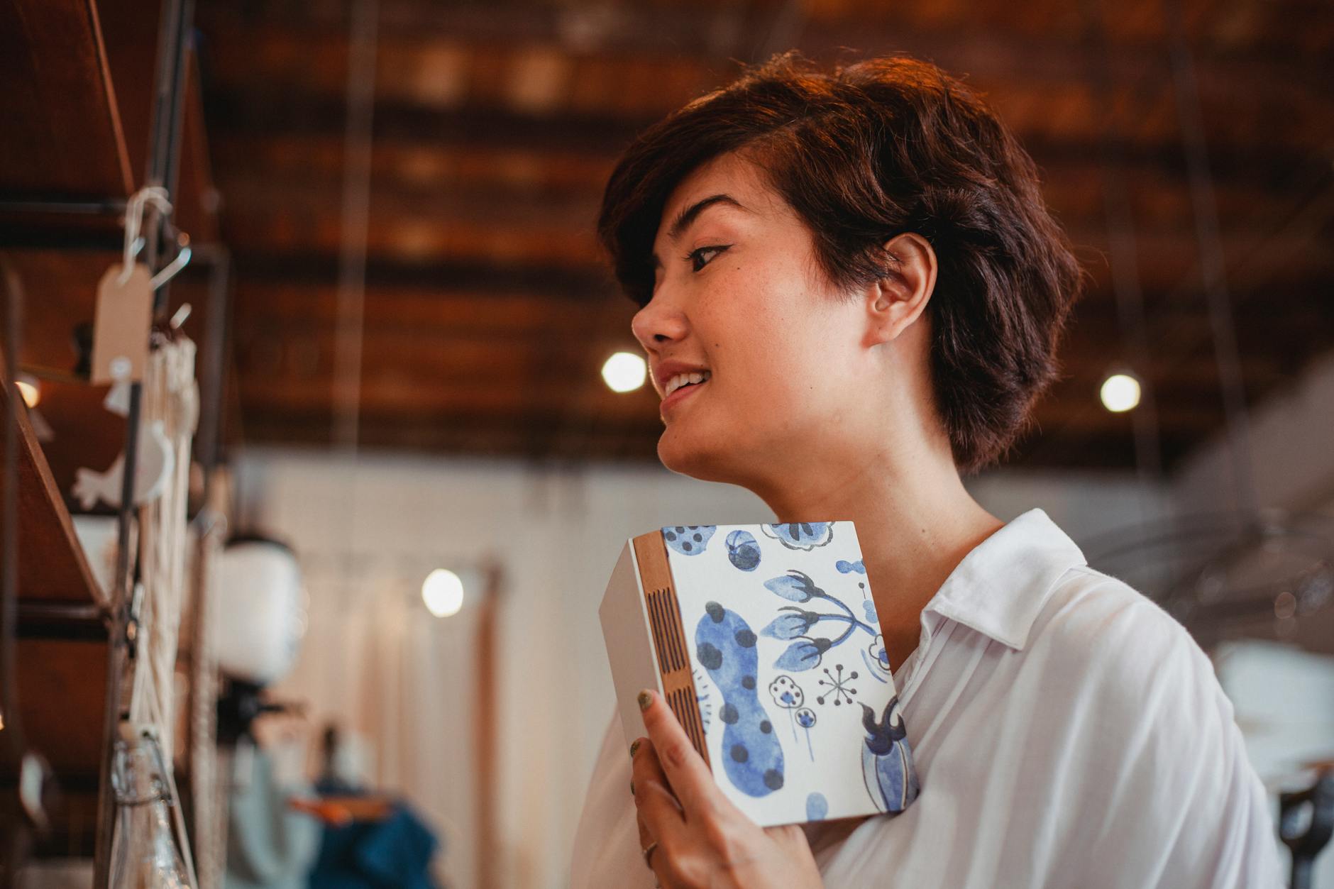 Side view of confident young Asian woman with short dark hair standing near shelf with various goods with stylish planner in hand in souvenir shop