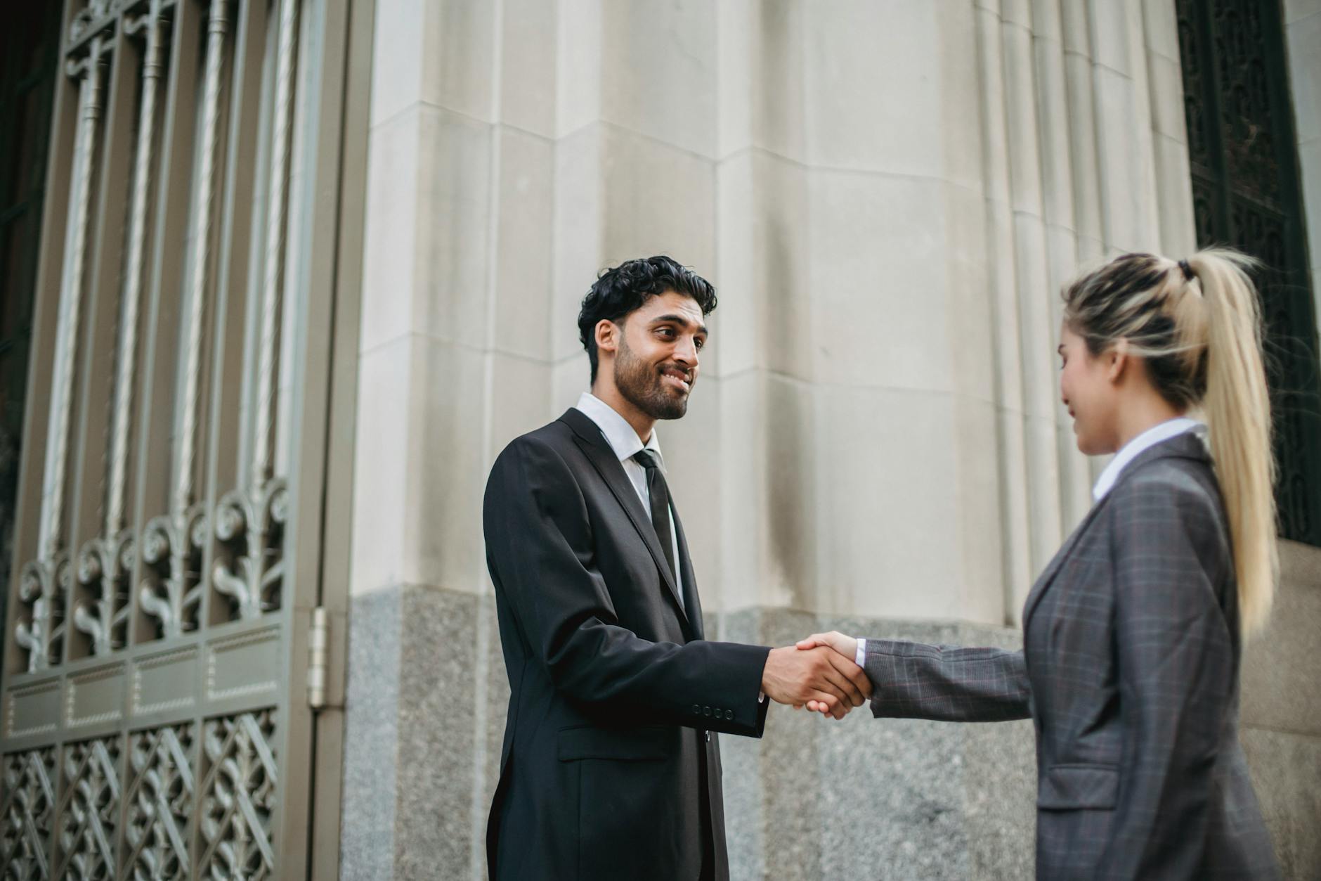 Two business professionals in formal attire shaking hands outside an elegant building, signifying partnership.