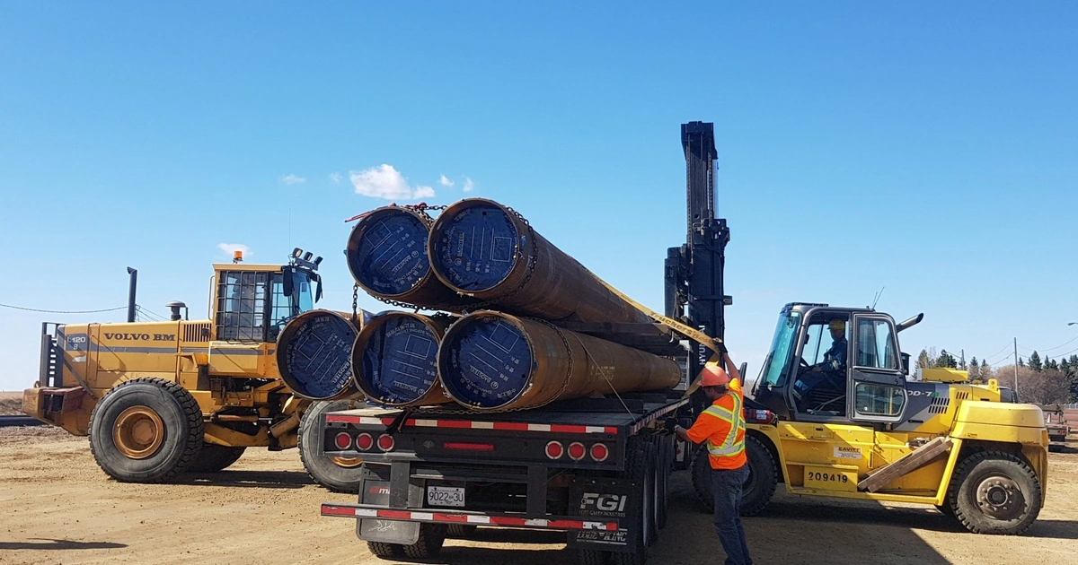 Heavy machinery loading steel pipe bundles at an Arrow Transportation reload terminal under clear skies.