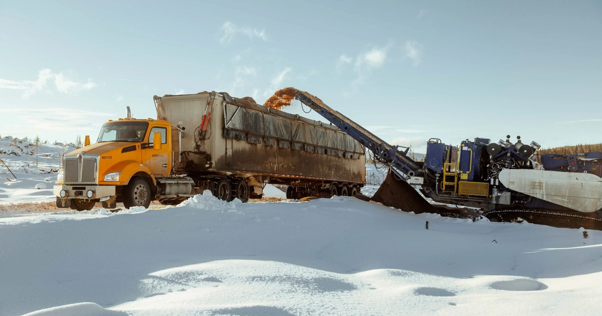 Yellow Arrow truck being loaded with processed wood fibre by a portable grinder in a snowy British Columbia landscape.