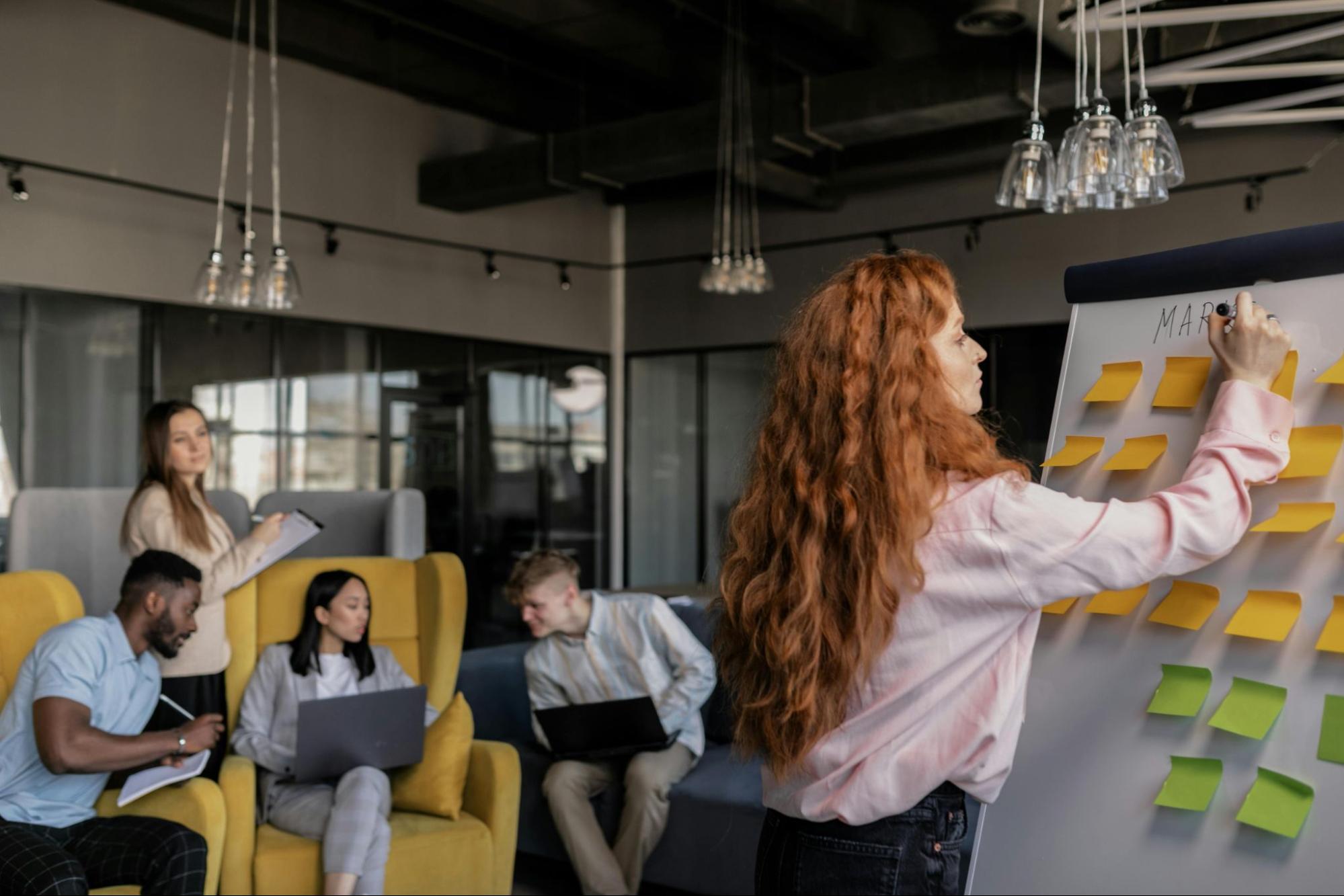 Red hair woman writing on a board in a marketing presentation while other people sitting behind discuss something.