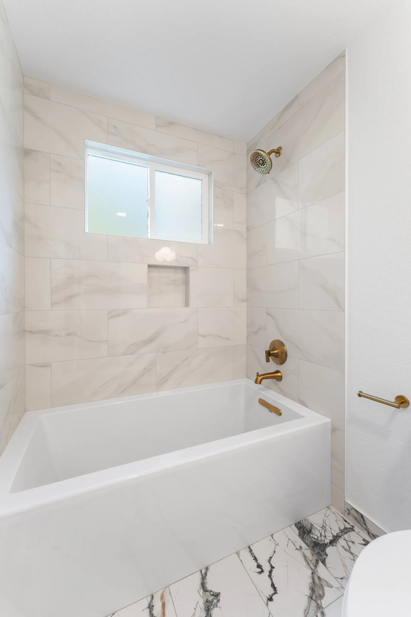 White tub surrounded by beige marble-patterned wall tiles installed horizontally with gold fixtures.