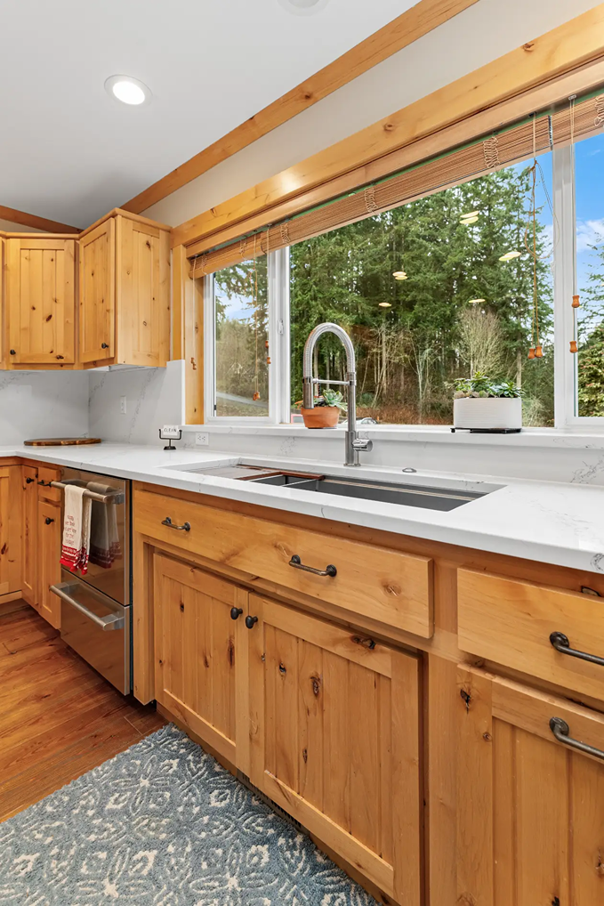 Kitchen sink area with white quartz countertops installed over natural pine cabinets